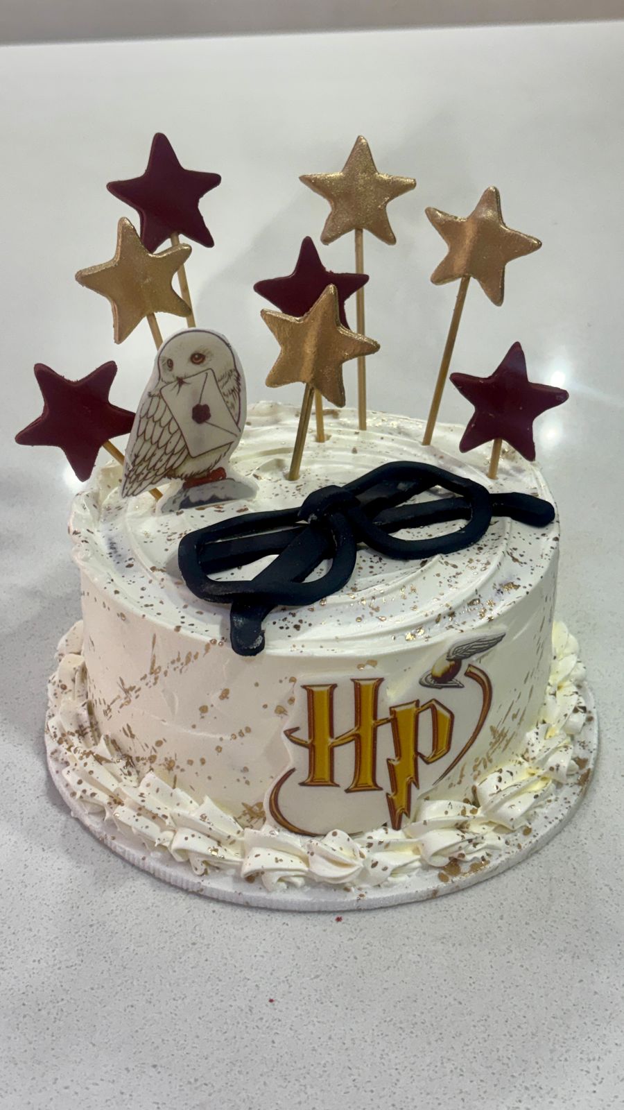 Torta blanca de Harry Potter con la búha Hedwig, lentes modelados en negro, estrellas doradas y granate, y salpicaduras de oro.