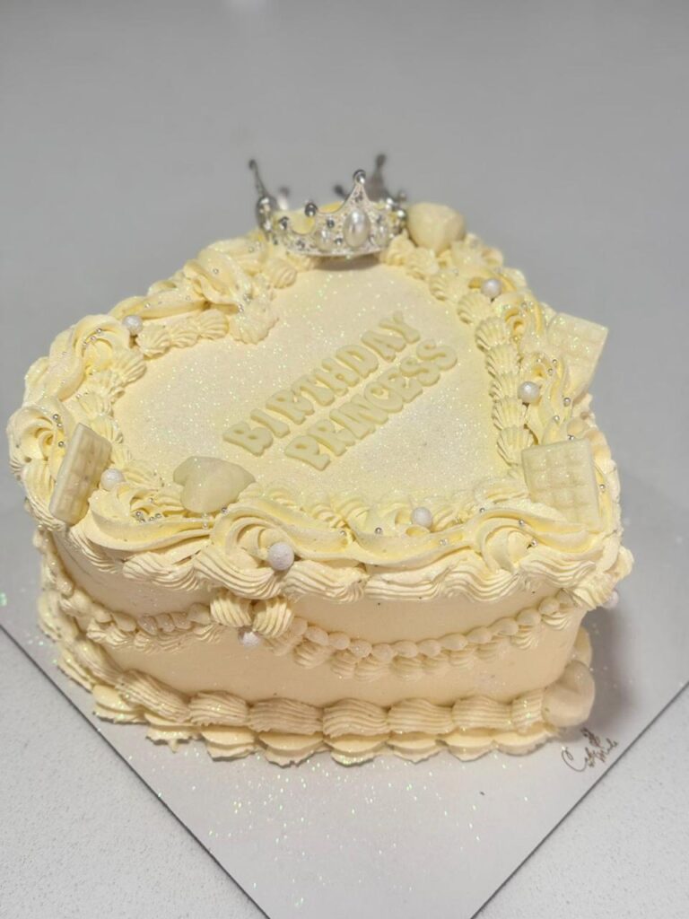 Torta corazón elegante con acabado marfil, detalles en crema texturizada y corona decorativa, ejemplo de diseños de pastelería premium para celebraciones exclusivas.
