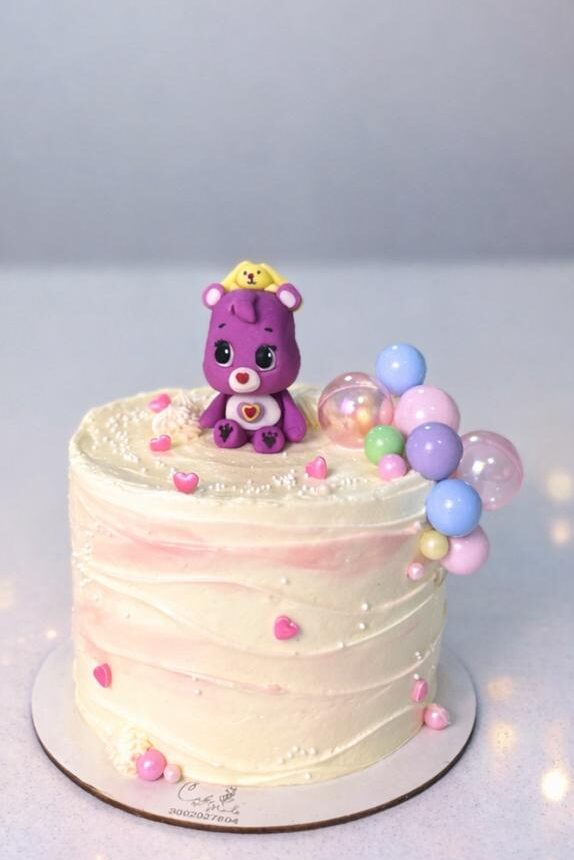 Torta artesanal blanca con degradado rosa suave, figura de Osito Cariñosito morado modelada a mano y racimo de burbujas de gelatina de colores pastel.