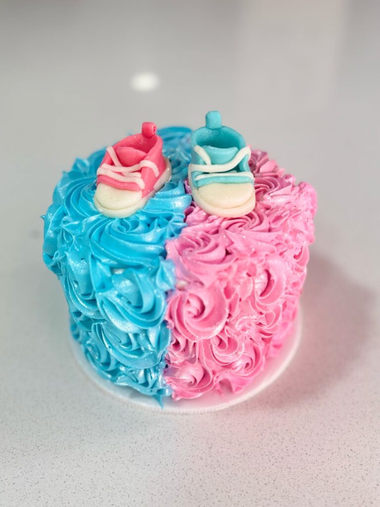 "Torta personalizada de baby shower con diseño mitad azul y mitad rosa, decorada con crema en forma de rosetones y zapatitos de fondant, ideal para revelar el género del bebé o celebrar la llegada de un nuevo integrante"