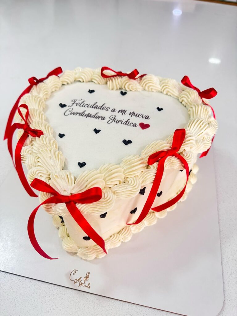 "Torta en forma de corazón decorada con crema blanca, lazos rojos y mensaje personalizado de felicitación para coordinadora jurídica, ideal para celebraciones profesionales y reconocimientos especiales"
