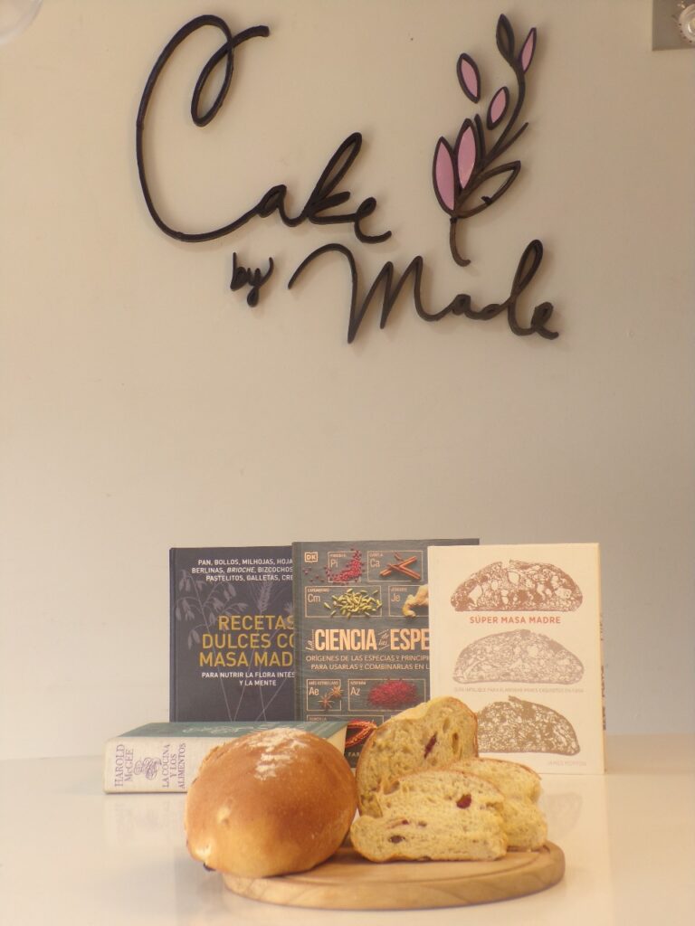 Pan artesanal elaborado con masa madre sobre tabla de madera, acompañado de libros sobre panadería saludable, frente al logo de Cake by Made en Bogotá.