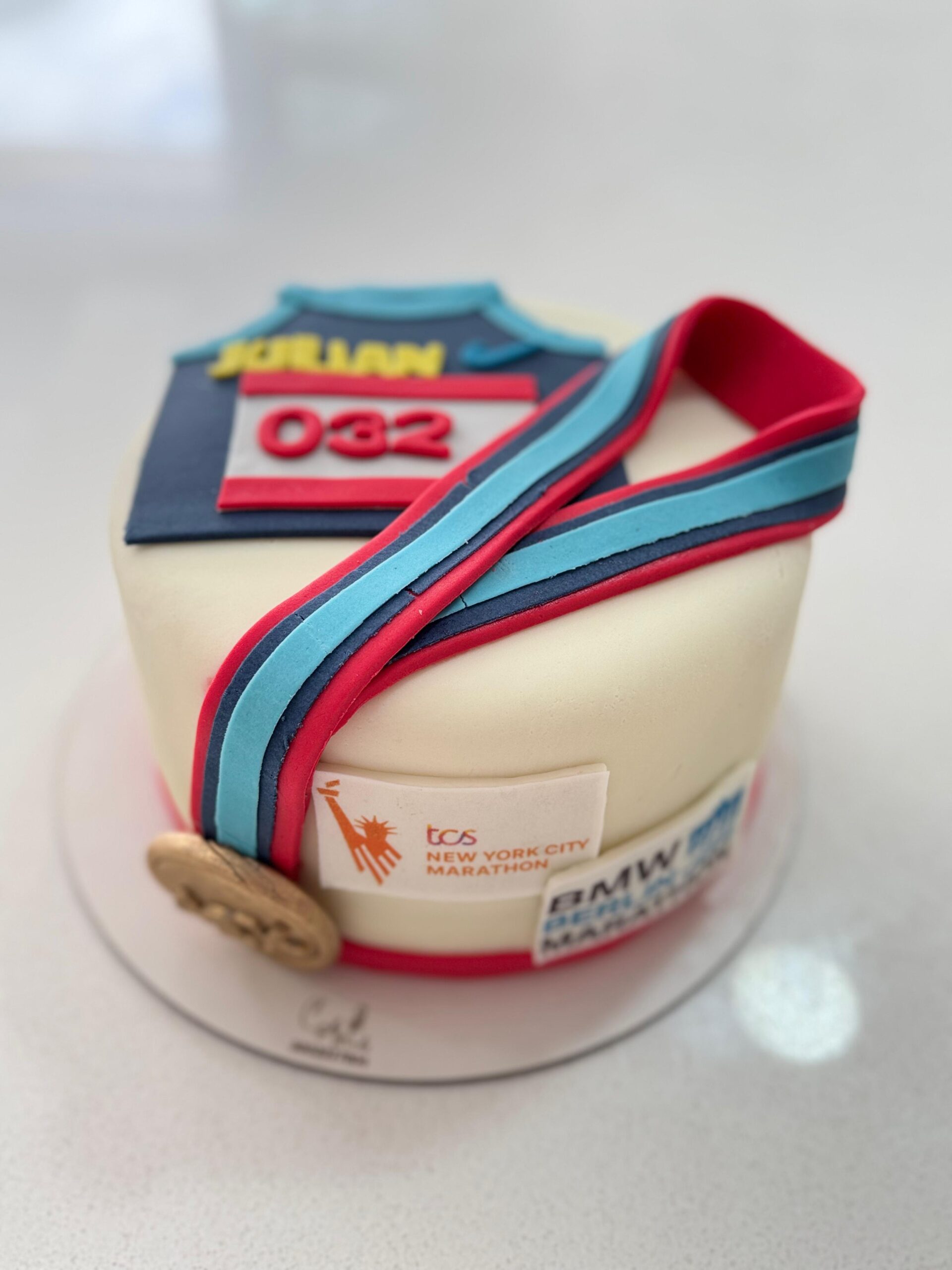 Pastel artesanal para corredores con temática de running, dorsal número 032 y detalles realistas de medalla y cinta de finisher.nombre "JOLIAN", y logos de la maratón de Nueva York (TCS New York City Marathon) y posiblemente de un patrocinador (BMW). El pastel está sobre una base redonda blanca.