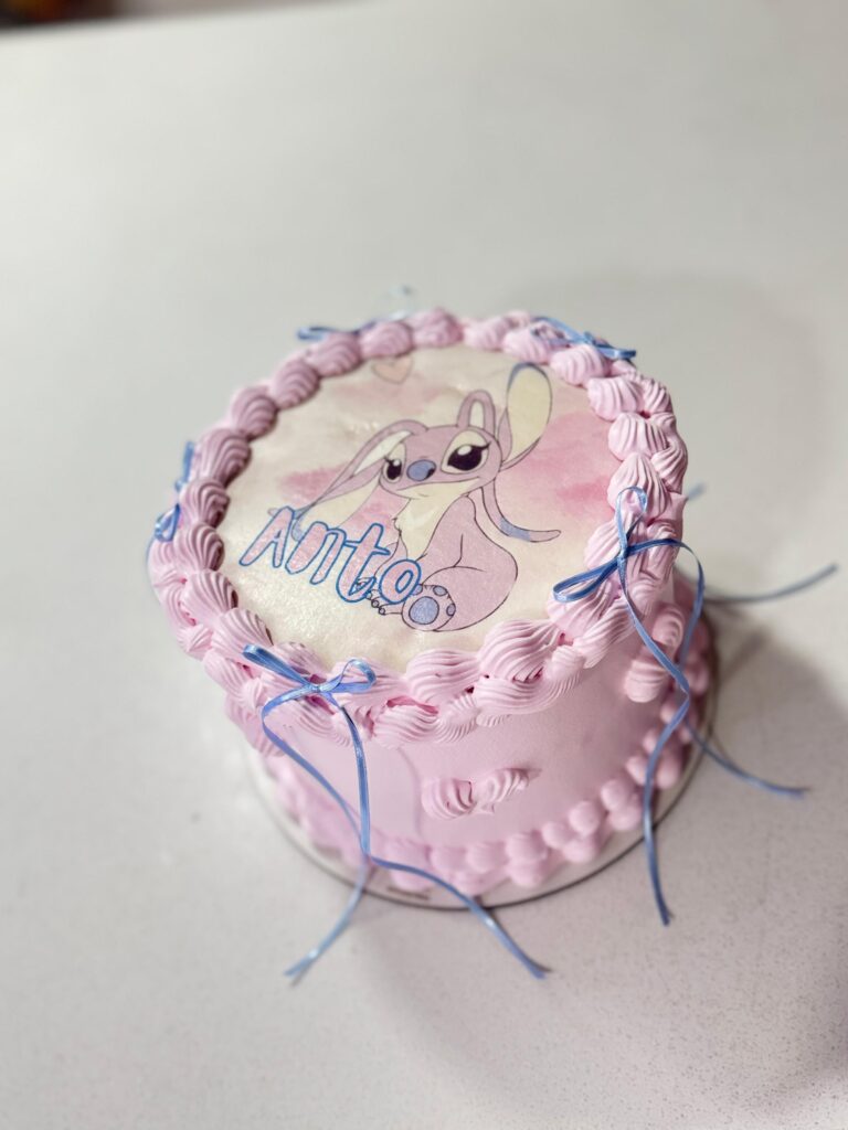 Torta temática de Angel, la adorable pareja de Stitch, decorada con crema rosa, cintas celestes y topper impreso comestible con nombre personalizado. Una opción dulce, tierna y única para cumpleaños infantiles o celebraciones especiales. Pastelería personalizada en Bogotá.