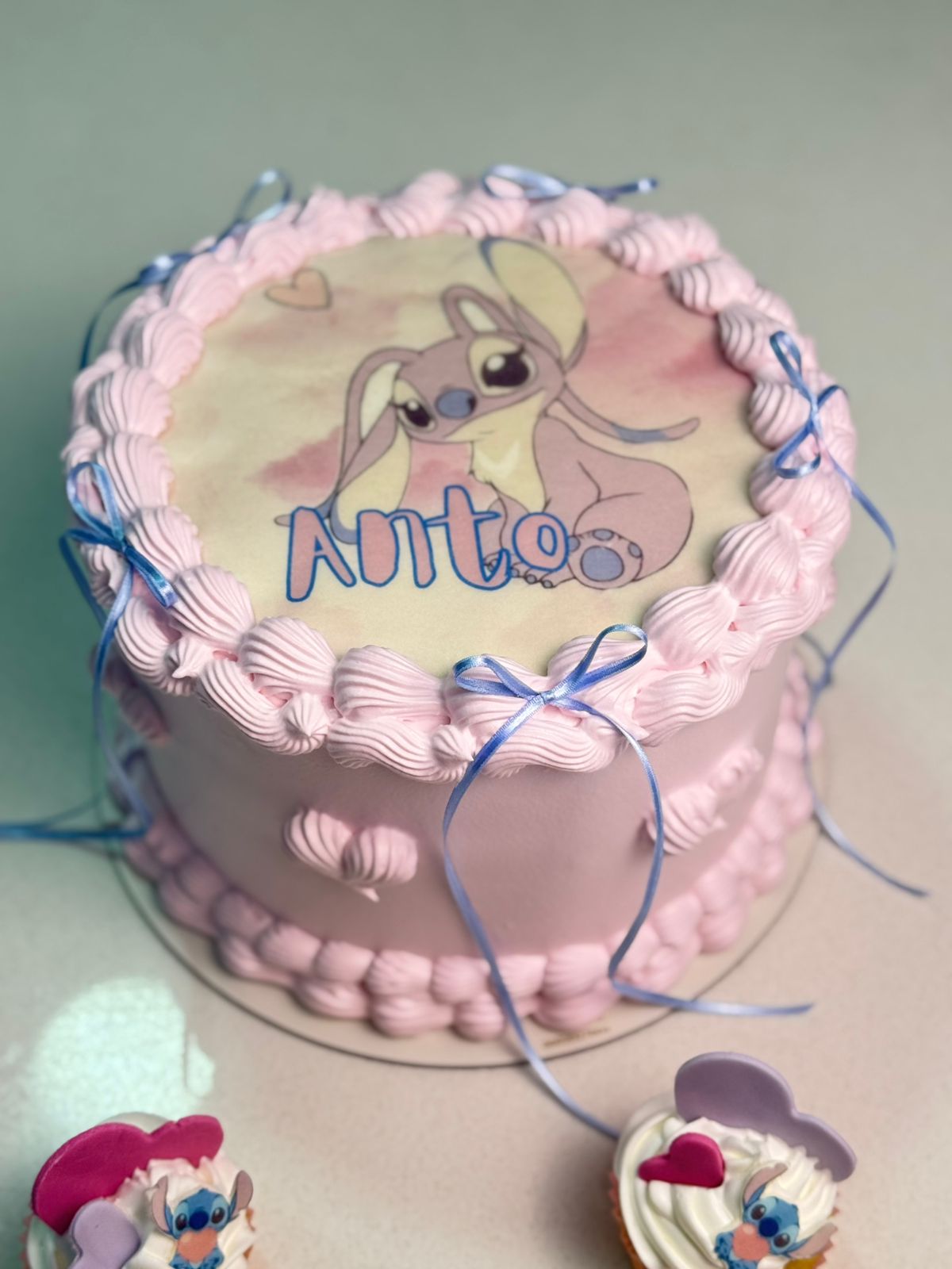 Torta básica personalizada Cake by Made con temática de Angel (Lilo & Stitch), frosting de buttercream rosa con técnica de boquilla rústica, moños celestes de cinta y oblea comestible central.